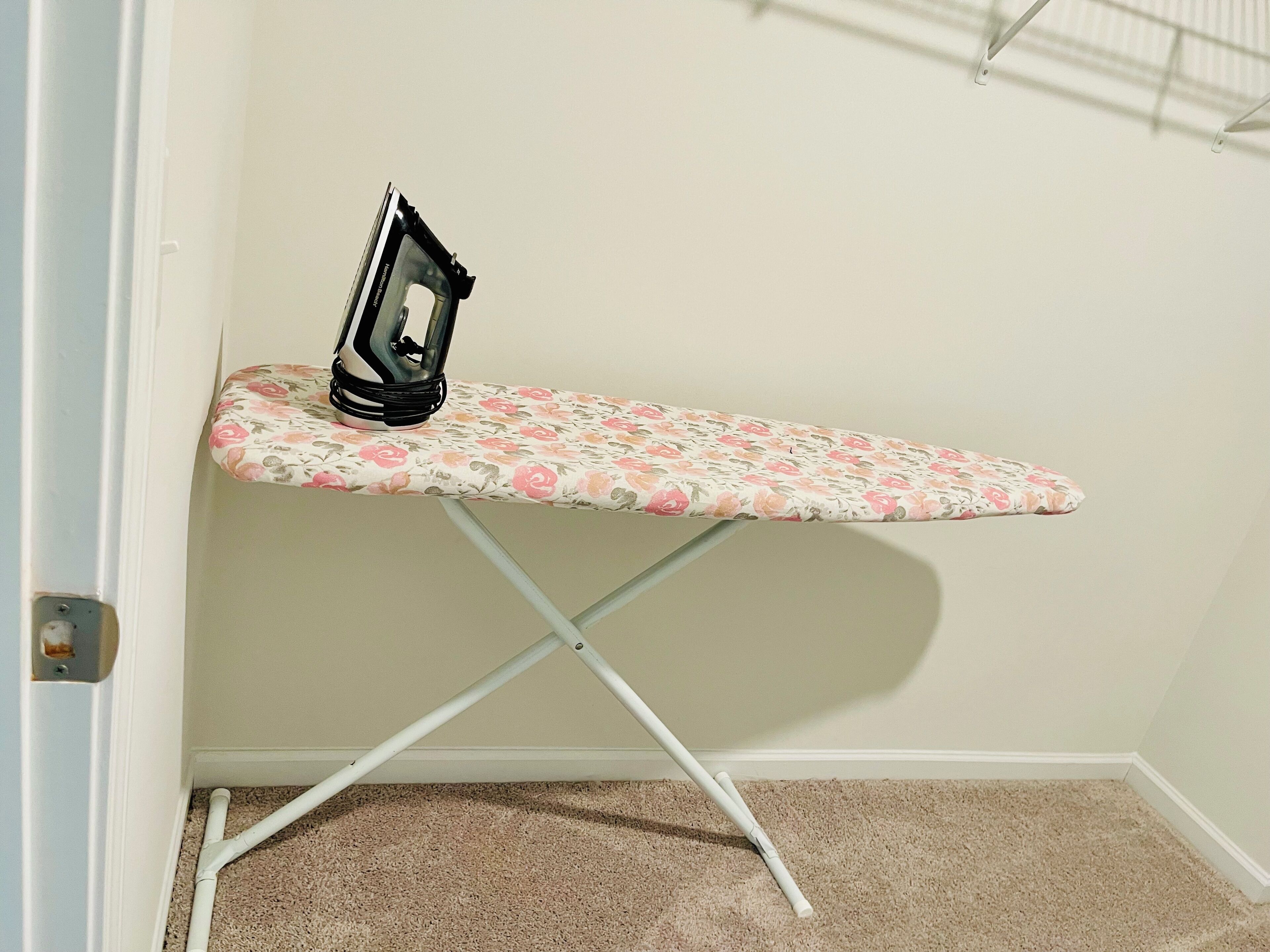 A corner ironing board with a floral pattern, a portable iron, and a white wall.