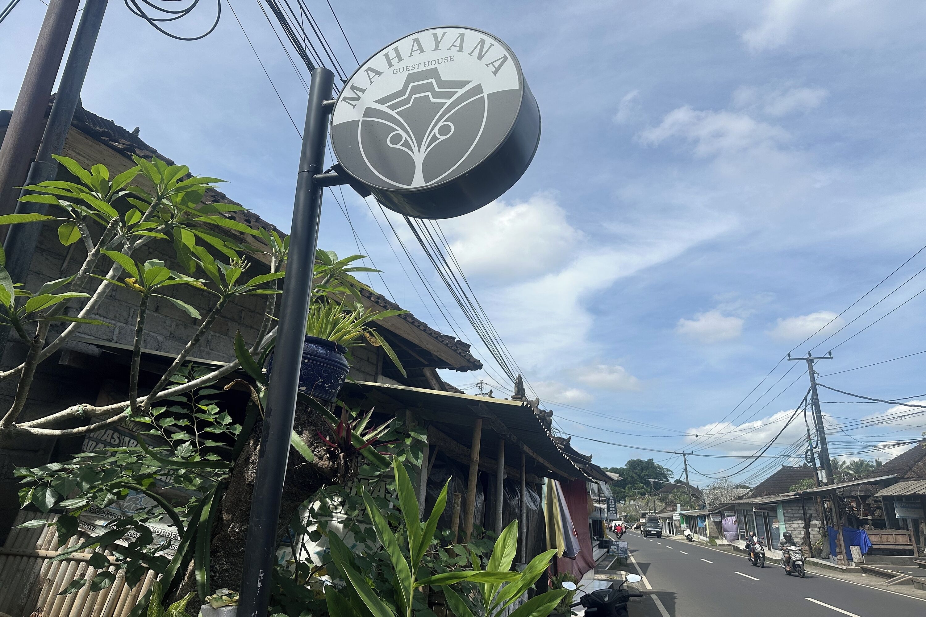 A sign for "MAHAYANA GUEST HOUSE" with a traditional design, located on a street with buildings and power lines in the background.