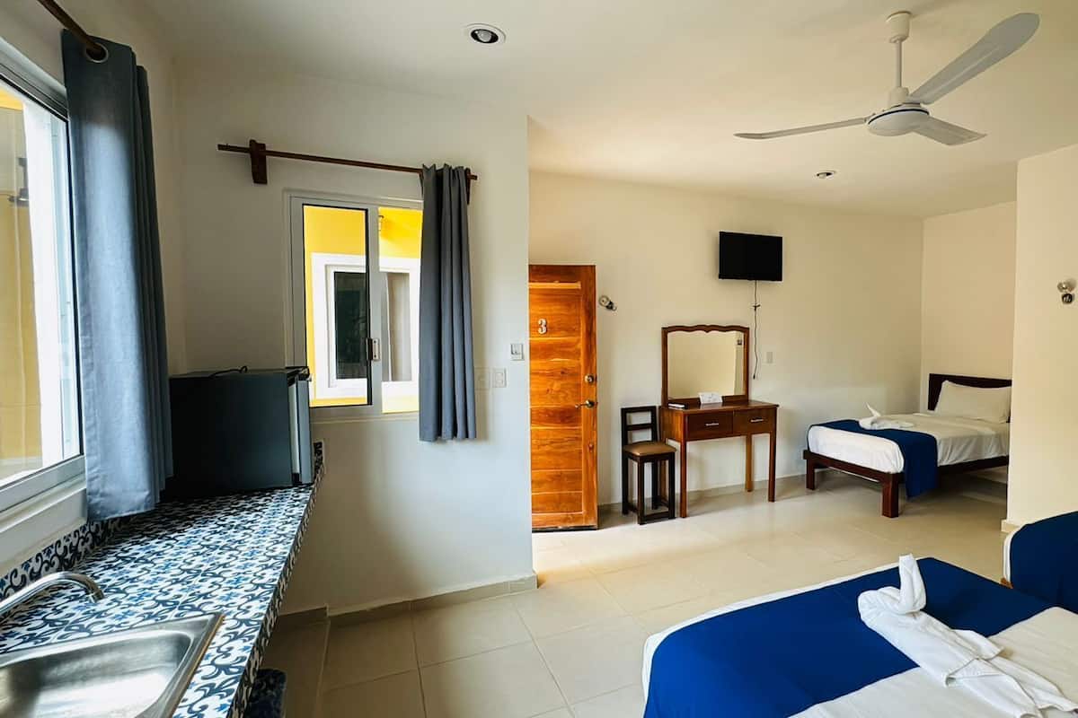 A hotel room with a kitchenette, a bed, a desk with a chair, a mirror, and a ceiling fan.