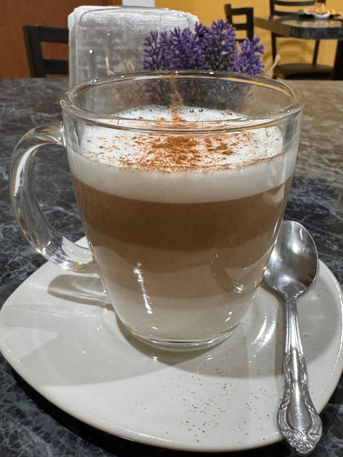 A clear glass cup of coffee with a layer of foam on top, placed on a white saucer with a spoon beside it.
