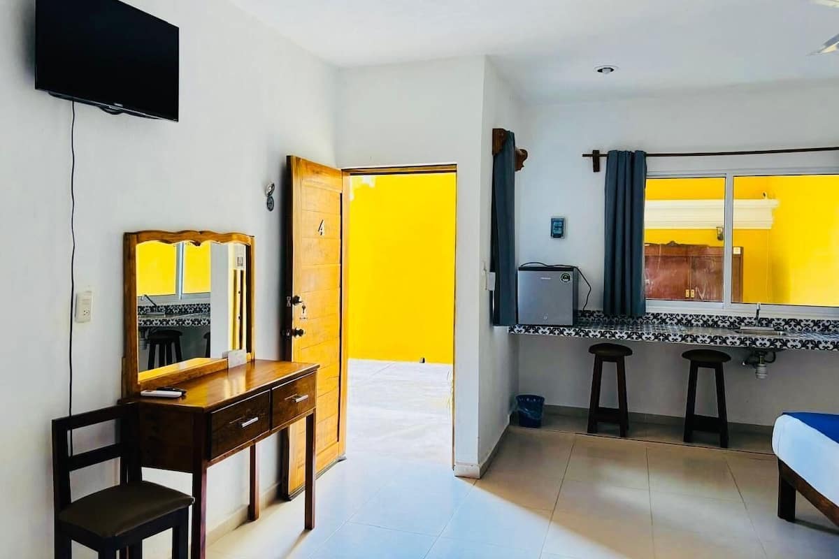 A hotel room with a TV, a desk with a mirror, a bed, a kitchen area with stools, and a bright yellow wall.