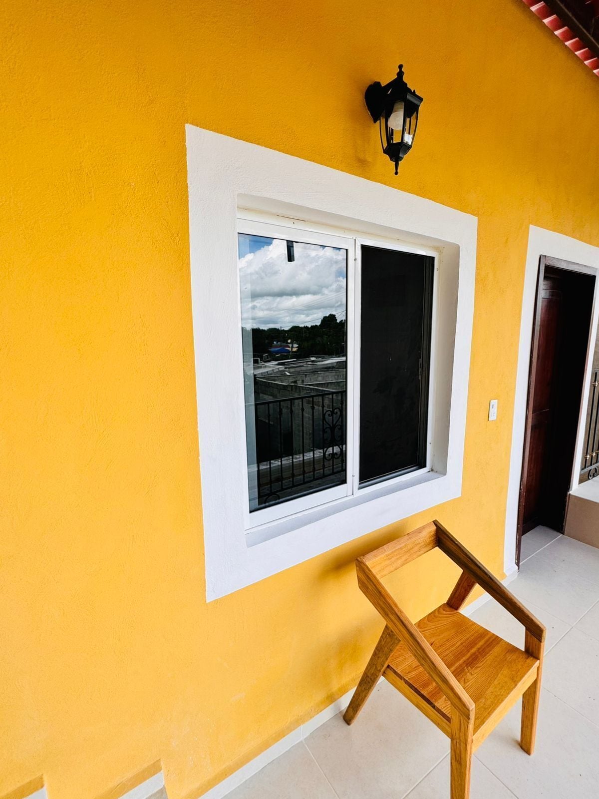 Una habitación con una pared amarilla, una ventana con vista a un edificio y una silla de madera.