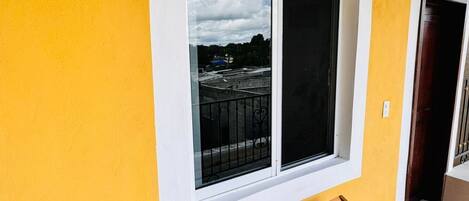 Una habitación con una pared amarilla, una ventana con vista a un edificio y una silla de madera.