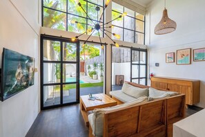 A spacious living area with a wooden sofa, a coffee table, and a hanging light fixture. Large windows provide natural light and views of a garden. - The Soho Loft by Pertama Stay (Pecatu)