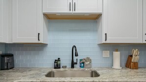 A kitchen with white cabinets, a blue tiled backsplash, a stainless steel sink, a coffee maker, a kettle, a dish soap bottle, a paper towel holder, and a knife block. - Sunny Haven #623: 1BR Townhome in Fort Walton (Fort Walton Beach)