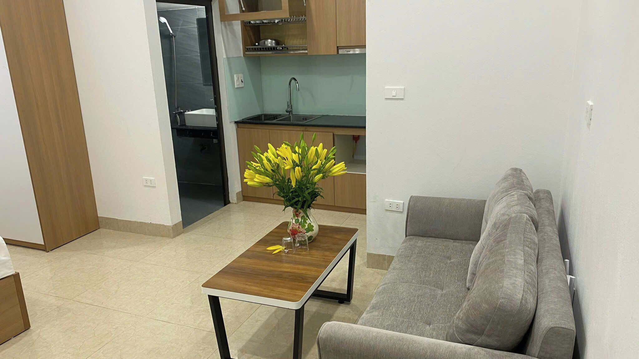 A compact living space with a kitchenette, a gray sofa, and a small wooden table with a vase of yellow flowers.