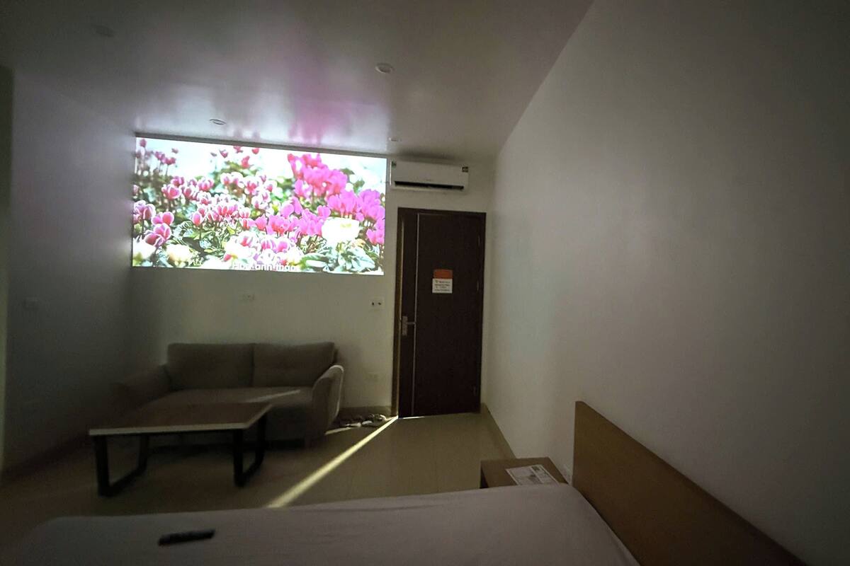 A small hotel room with a bed, a sofa, a table, and a window with pink flowers.