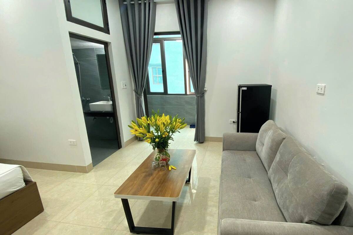 A modern living room with a sofa, a wooden coffee table with a vase of flowers, a TV, and a view of the outside through a window with curtains.