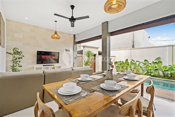 Un comedor moderno con una mesa de madera preparada para cuatro personas, un sofá y un televisor mostrando 'NETFLIX'.