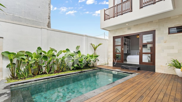 A swimming pool with a wooden deck, surrounded by lush greenery and a modern building with a balcony. - Cavara Villa by Kozystay - Ungasan (Ungasan)