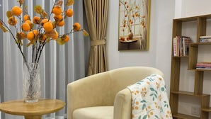A living room with a beige sofa, a round wooden table with a vase, a patterned throw, a bookshelf, and a geometric patterned floor. - Grand C Homestay (Hanoi)