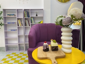 A purple sofa, a yellow table with a wooden board, and a white vase with flowers.