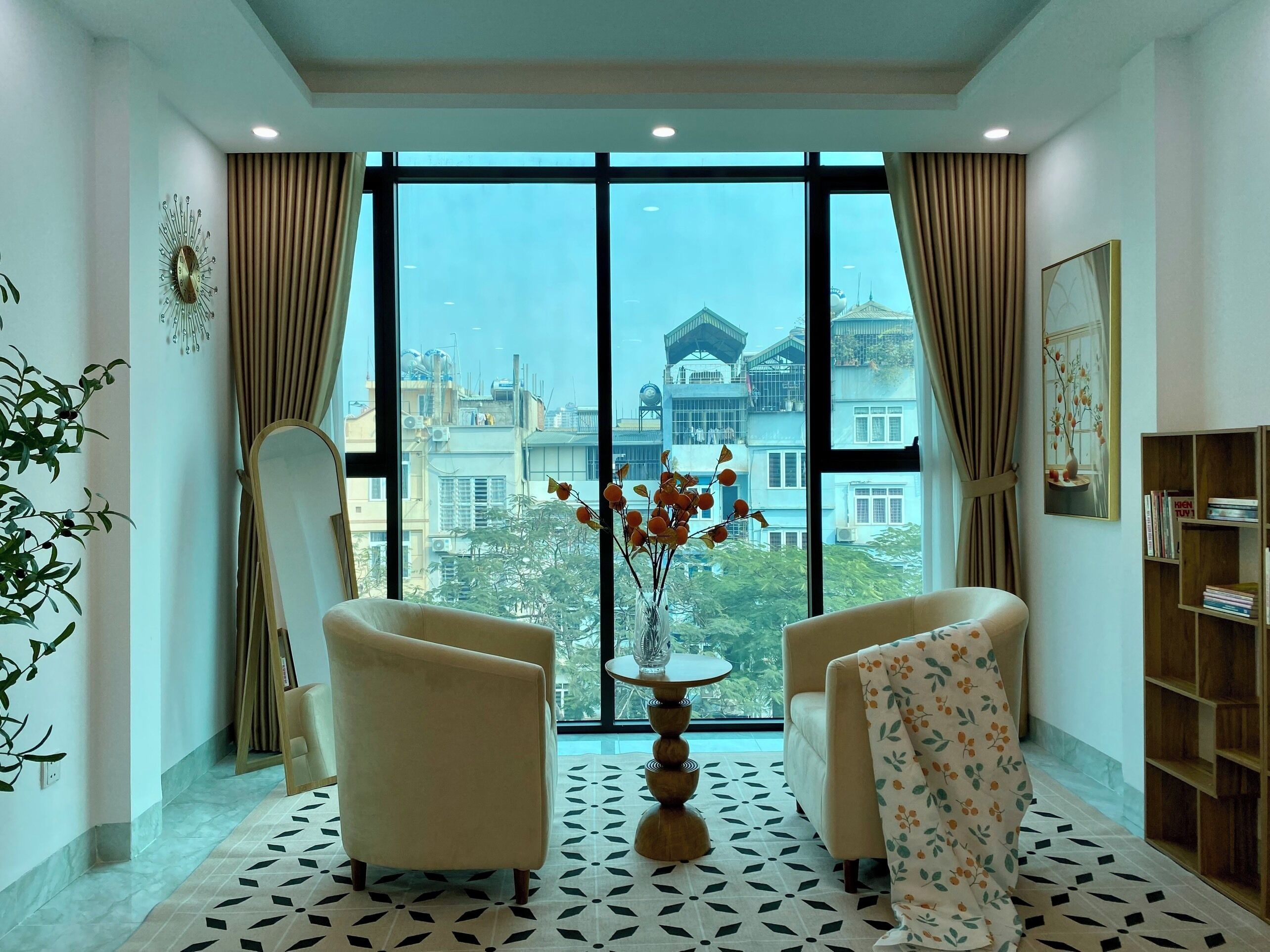 A room with two armchairs, a round table, and a view of buildings through large windows.
