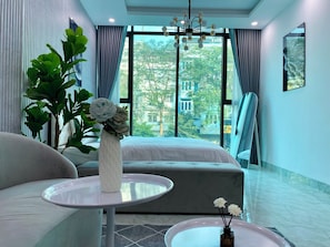 A modern hotel room with a large window, a bed, a sofa, a small table, and a potted plant. - Grand C Homestay (Hanoi)