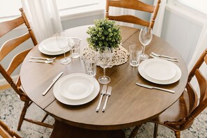 Une table de salle à manger ronde en bois, dressée avec de l’assiette blanche, des couverts et des verres transparents, et agrémentée d’une petite plante verte au centre.