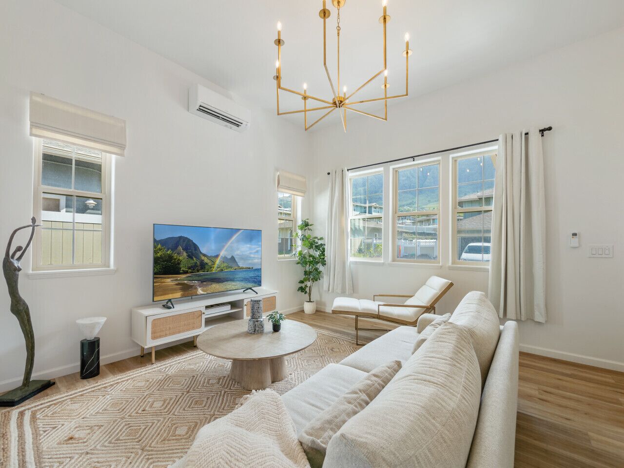A spacious living room with a large flat-screen TV, a modern chandelier, and a large window with curtains.
