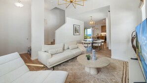 A spacious living room with a white sofa, a round coffee table, and a chandelier. There is a staircase leading to another level, and a dining area in the background. - Ocean Views|Outdoor Kids Play Set|BBQ|Beach 6 Min (Waianae)