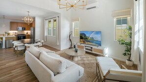 A modern living room with a sofa, coffee table, and TV. There is a kitchen in the background with a dining area. - Ocean Views|Outdoor Kids Play Set|BBQ|Beach 6 Min (Waianae)