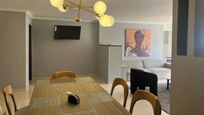 Ein Essbereich mit einem langen Tisch, Stühlen, einer Deckenleuchte, einem Flachbildfernseher und einer Couch im Hintergrund.