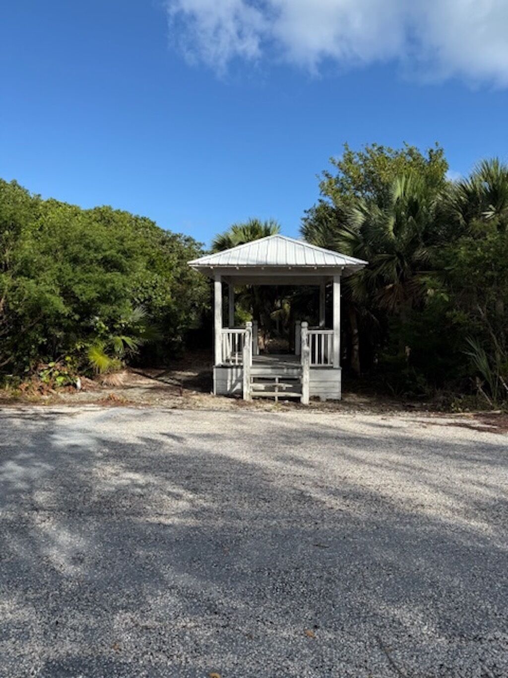 A gazebo with a gravel driveway leading to it, surrounded by lush greenery and palm trees.