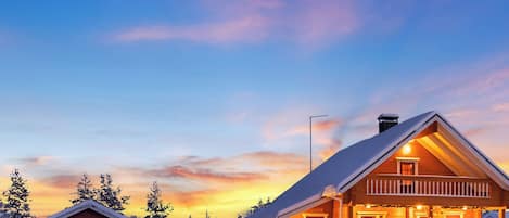 A wooden cabin with a balcony, surrounded by snow-covered trees, during sunset.