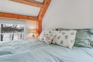 Een slaapkamer met een bed, kussens, een nachtkastje, een lamp en een raam met uitzicht op bomen.