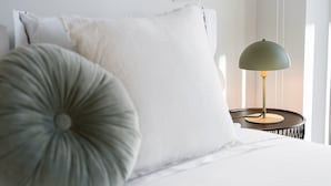 Uma cama bem arrumada com roupa de cama branca, um travesseiro cinzento e uma luminária verde sobre uma mesa de cabeceira.
