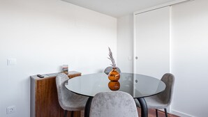 Uma sala de jantar moderna com mesa de vidro, duas cadeiras cinzas estofadas, um aparador de madeira e um vaso com plantas secas.