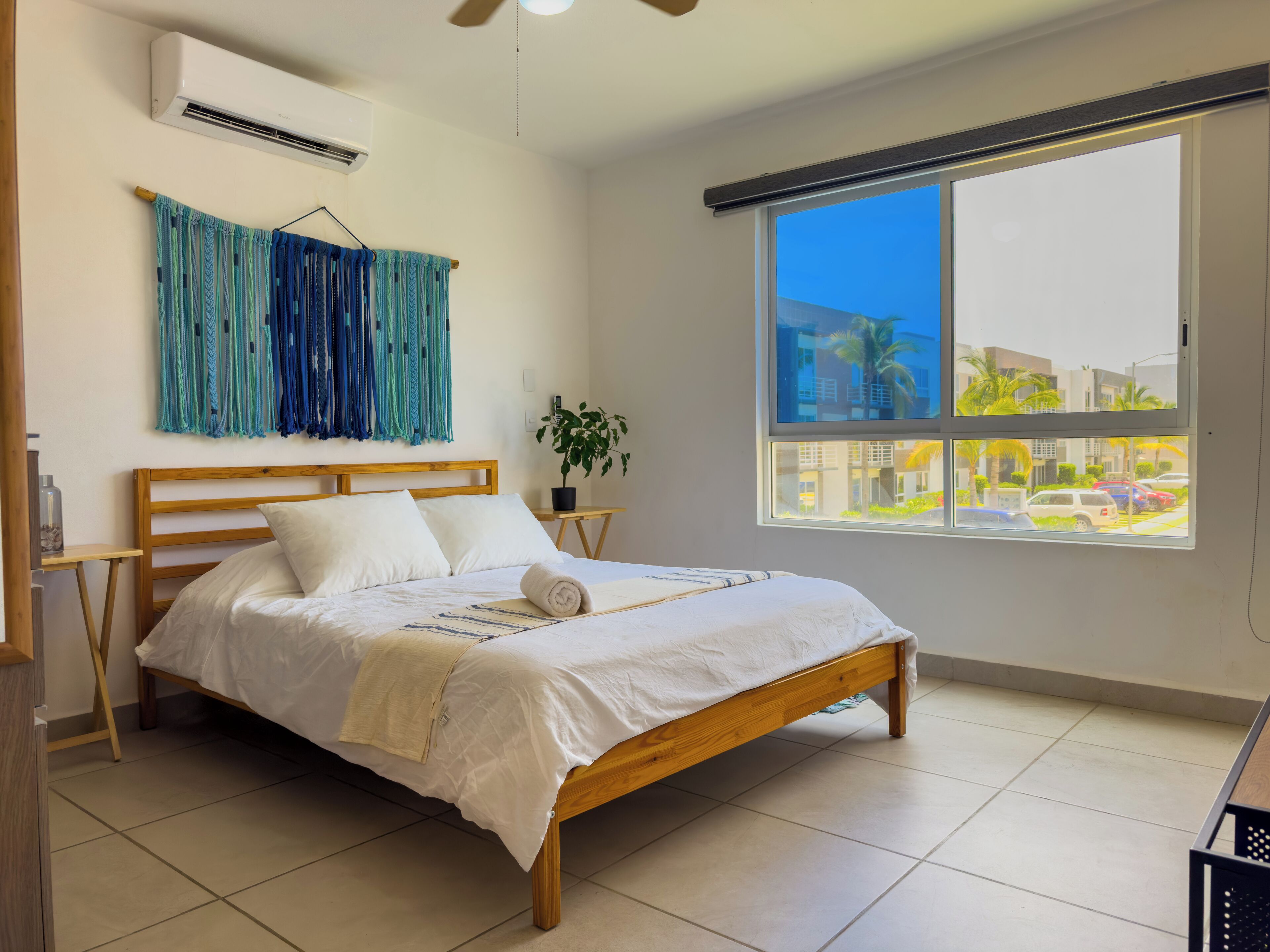 A bedroom with a wooden bed, white bedding, a large window with a view of buildings and cars, and a ceiling fan.