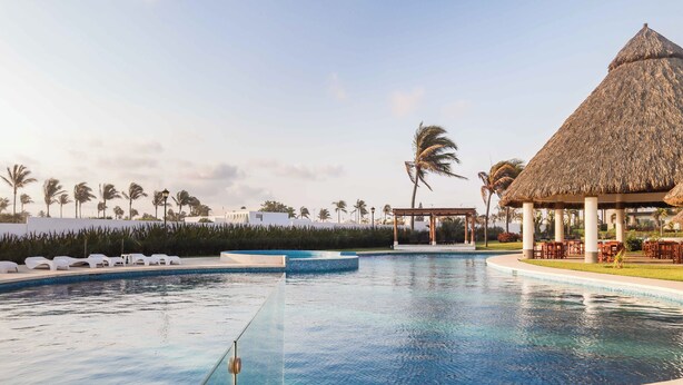Una piscina di un resort con lettini, un padiglione con tetto di paglia e palme.