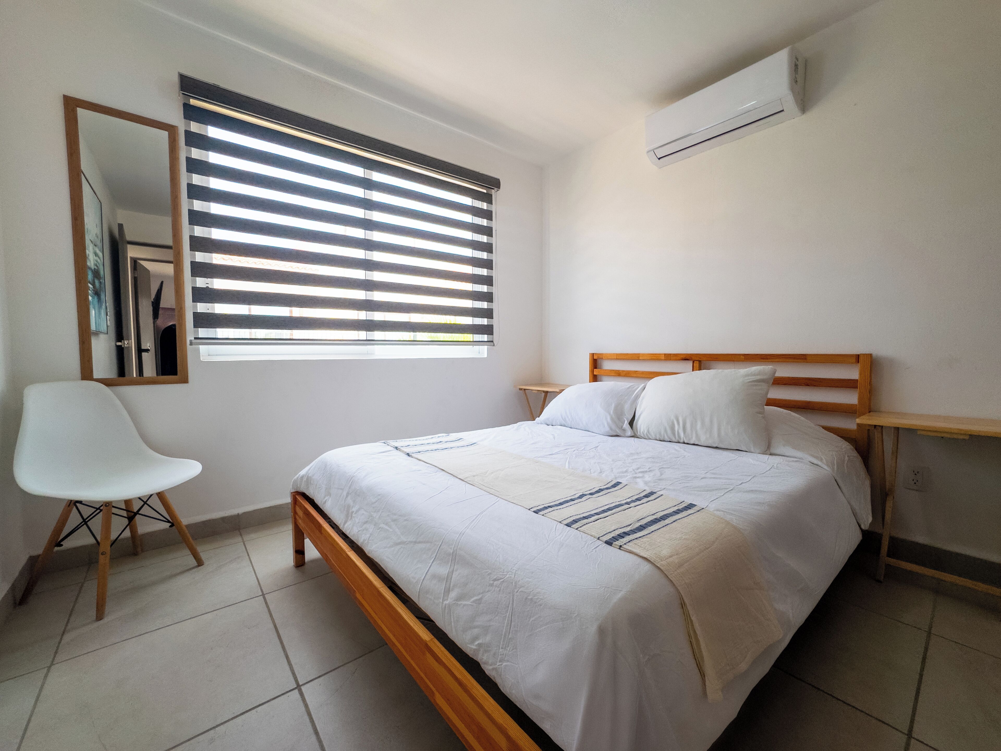 A bedroom with a bed, a chair, a window with blinds, and an air conditioning unit.