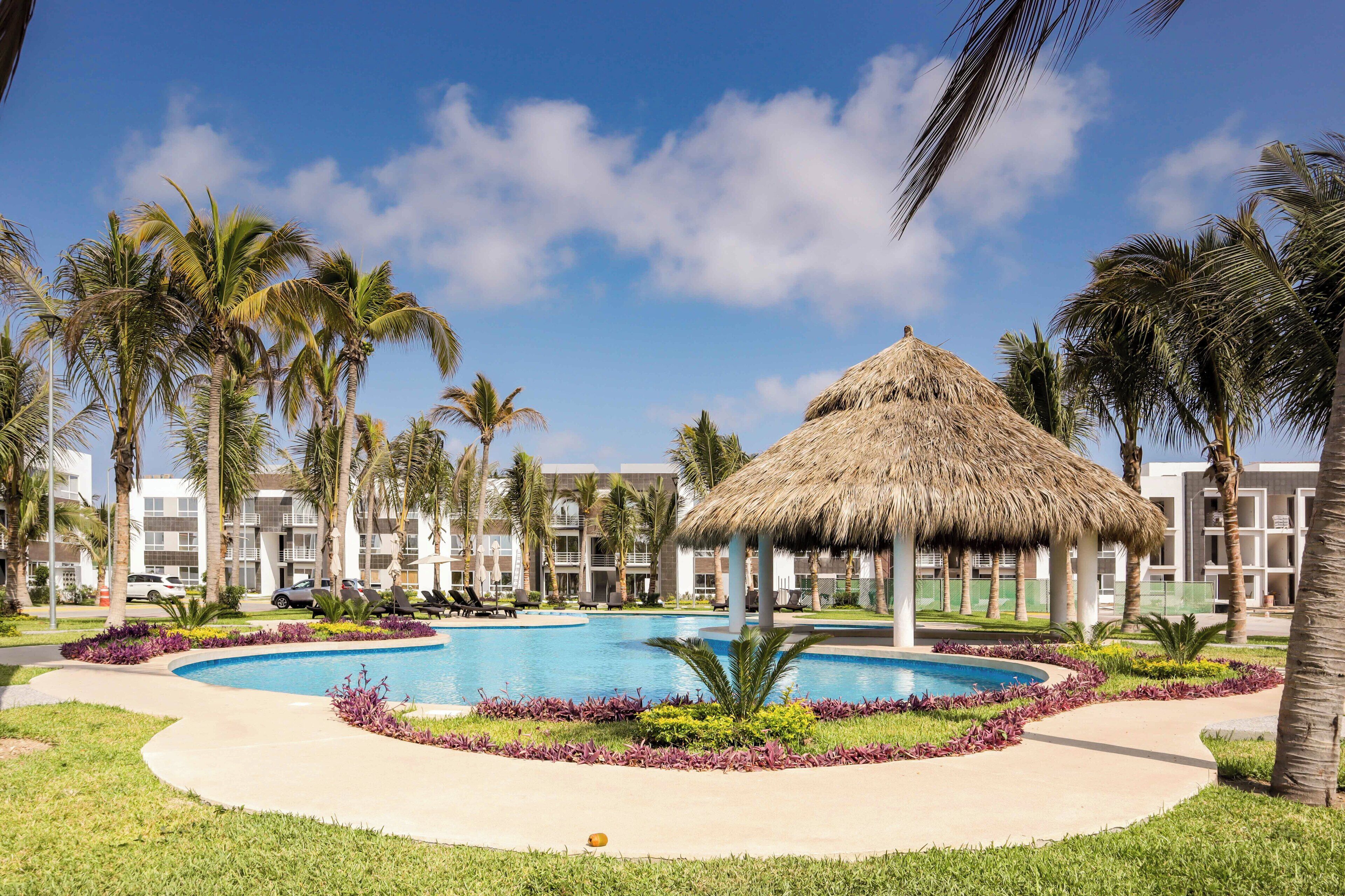 Zwembadgedeelte van een resort met een paviljoen met rieten dak, palmbomen en een zwembad omringd door een aangelegde tuin.