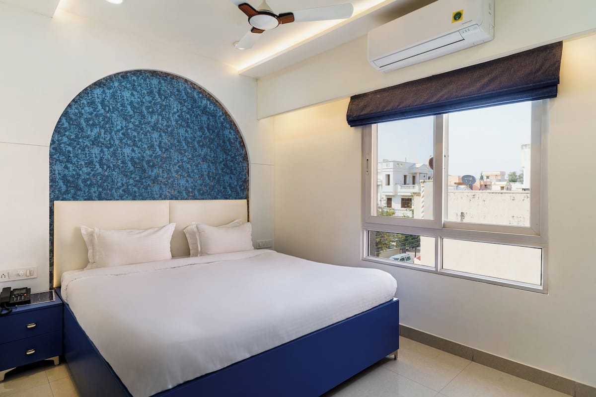 Une chambre d’hôtel moderne avec un grand lit, une tête de lit bleue, une fenêtre donnant sur des immeubles et un appareil de climatisation.