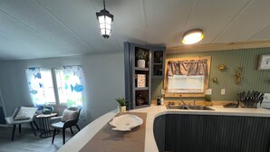 A kitchen with a white countertop, a sink, and a window with blinds. There are chairs and a table in the adjacent dining area. - Charming 2-bedroom mobile home with WiFi and AC in Okeechobee (Okeechobee)
