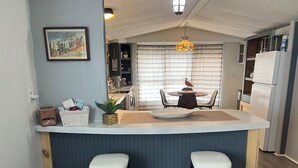 A compact kitchen with a central island, two stools, a dining table with chairs, a hanging light fixture, and a refrigerator. - Charming 2-bedroom mobile home with WiFi and AC in Okeechobee (Okeechobee)