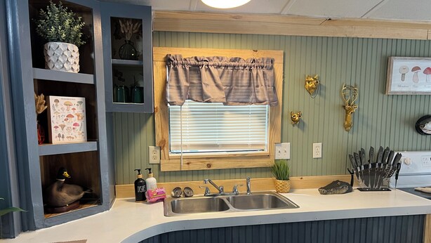 A kitchen with a built-in sink, wooden shelves, a window with blinds, and a knife block. - Charming 2-bedroom mobile home with WiFi and AC in Okeechobee (Okeechobee)