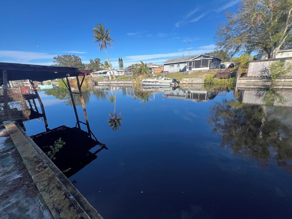 A calm water body reflecting buildings and trees, with a dock on the left and a house with a porch on the right.