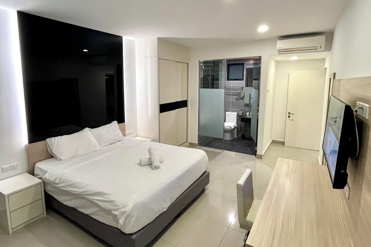 Una habitación de hotel moderna con una cama grande, mesitas de noche, un televisor y un baño visible a través de un panel de vidrio.