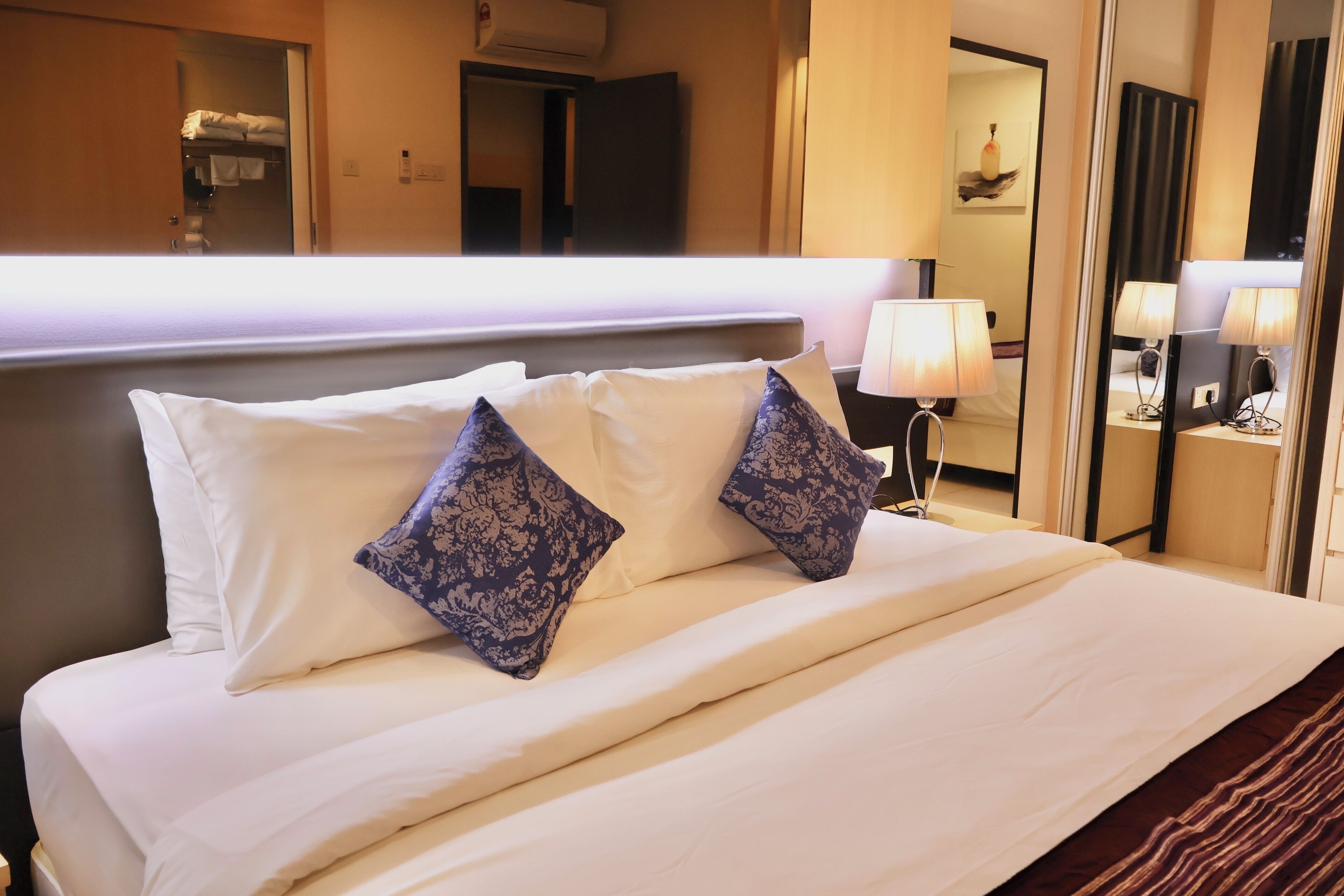 A hotel room with a neatly made bed, a bedside lamp, and a mirror reflecting the room's interior.