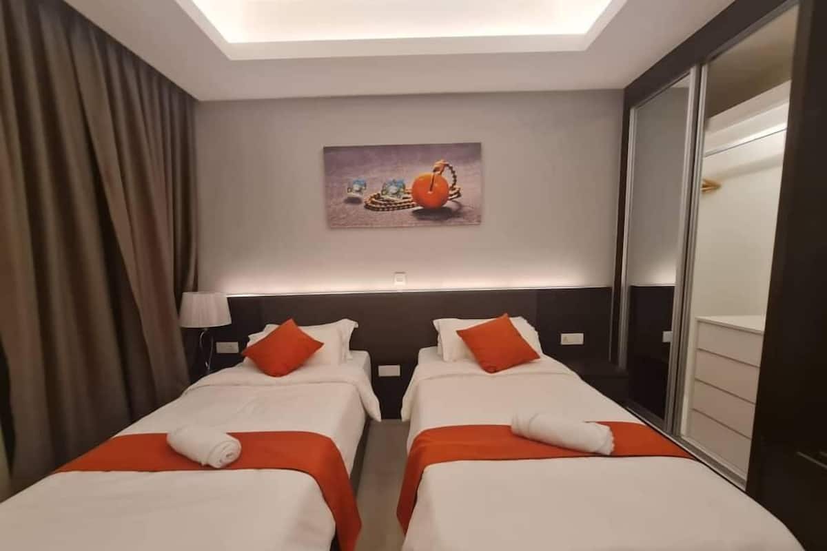 Habitación de hotel con dos camas, cada una con ropa de cama blanca y detalles en naranja, una lámpara de techo, un cuadro en la pared y un espejo grande.