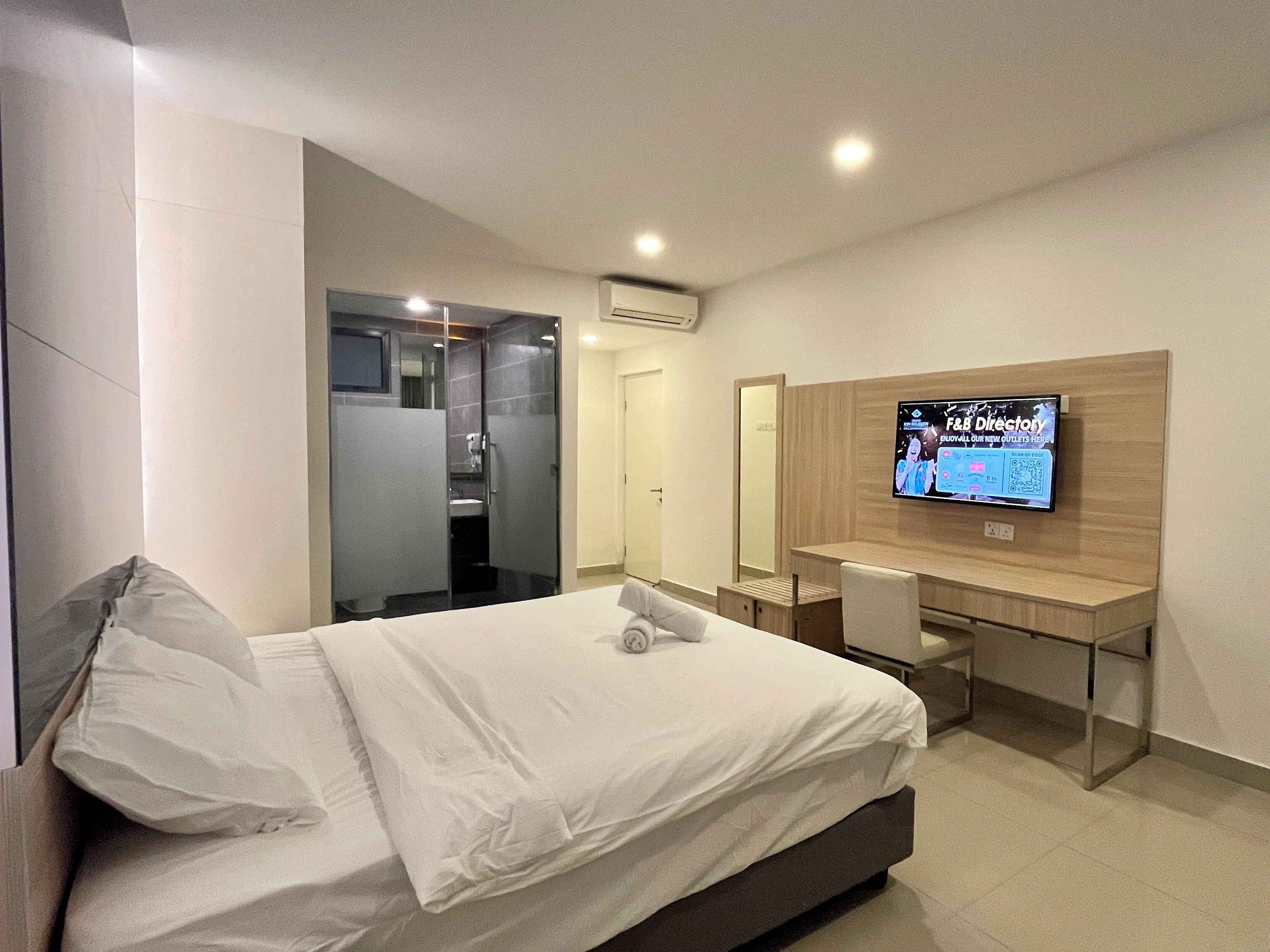 A hotel room with a bed, a desk, a TV, and a bathroom visible through a glass door.