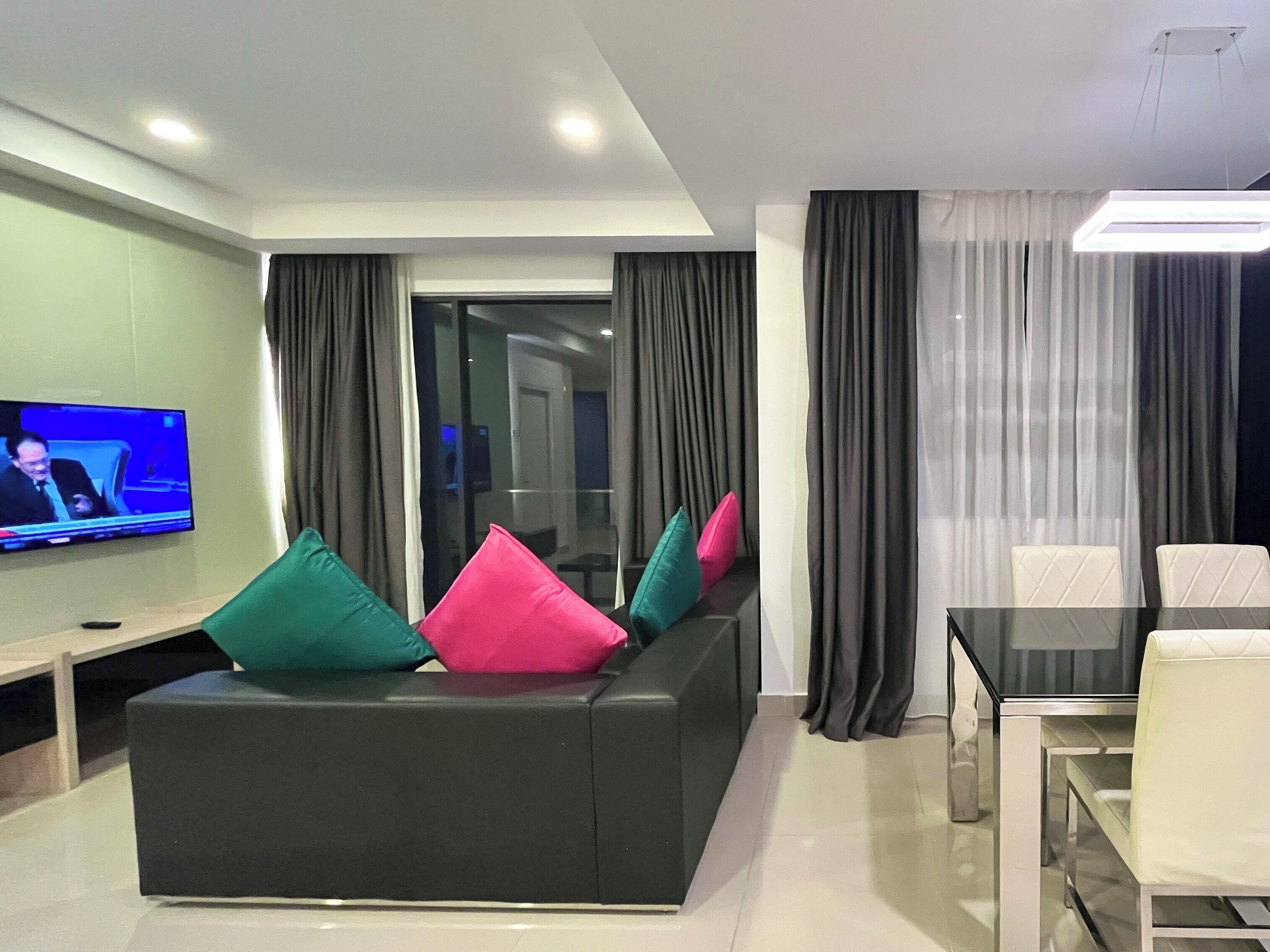 A modern living room with a black sofa, colorful cushions, a flat-screen TV, a glass dining table, and a view into the adjacent room.
