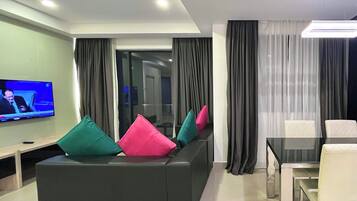 A modern living room with a black sofa, colorful cushions, a flat-screen TV, a glass dining table, and a view into the adjacent room.