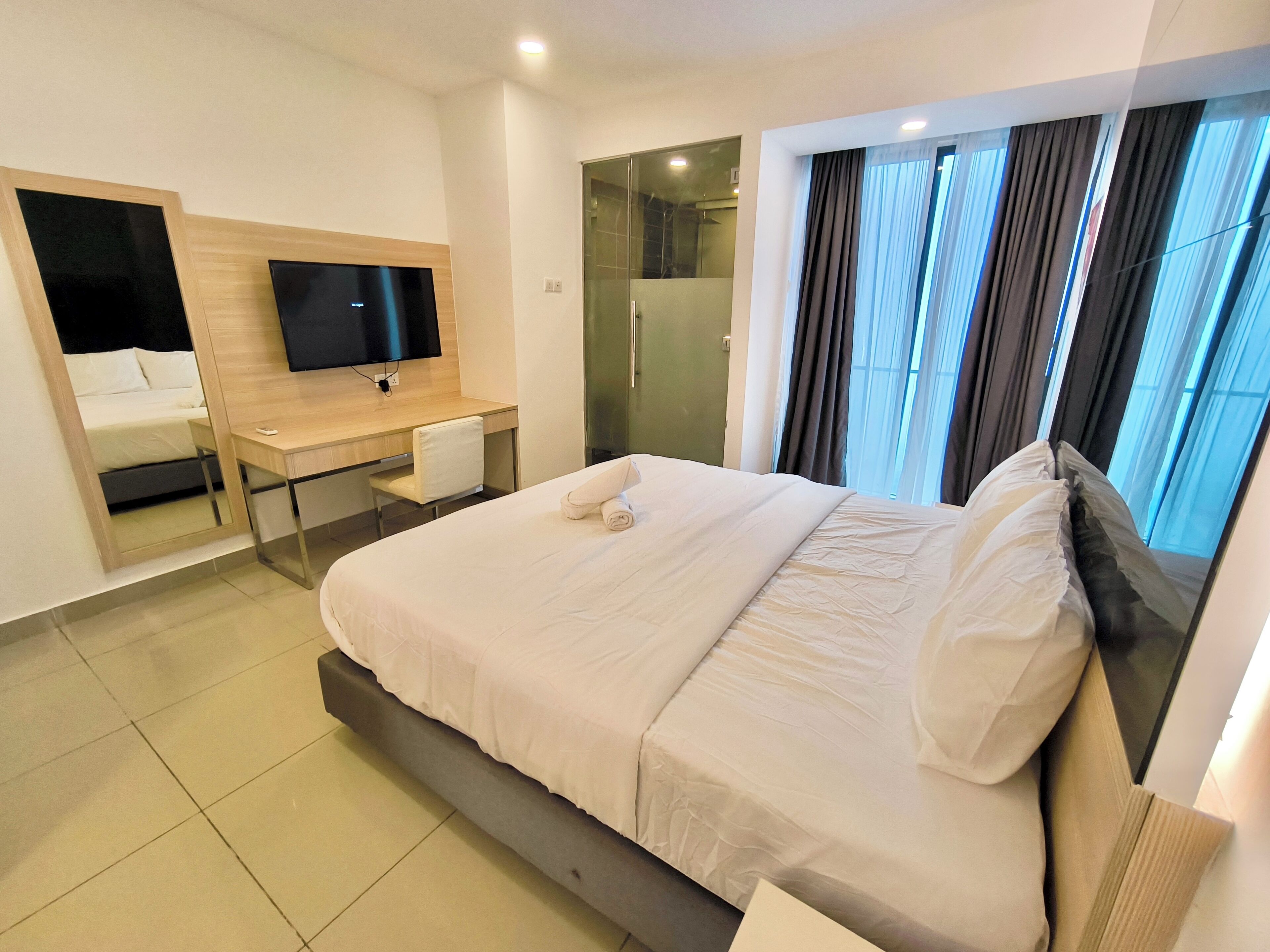 A modern hotel room with a large bed, a desk with a chair, a TV, and a bathroom with a glass door.