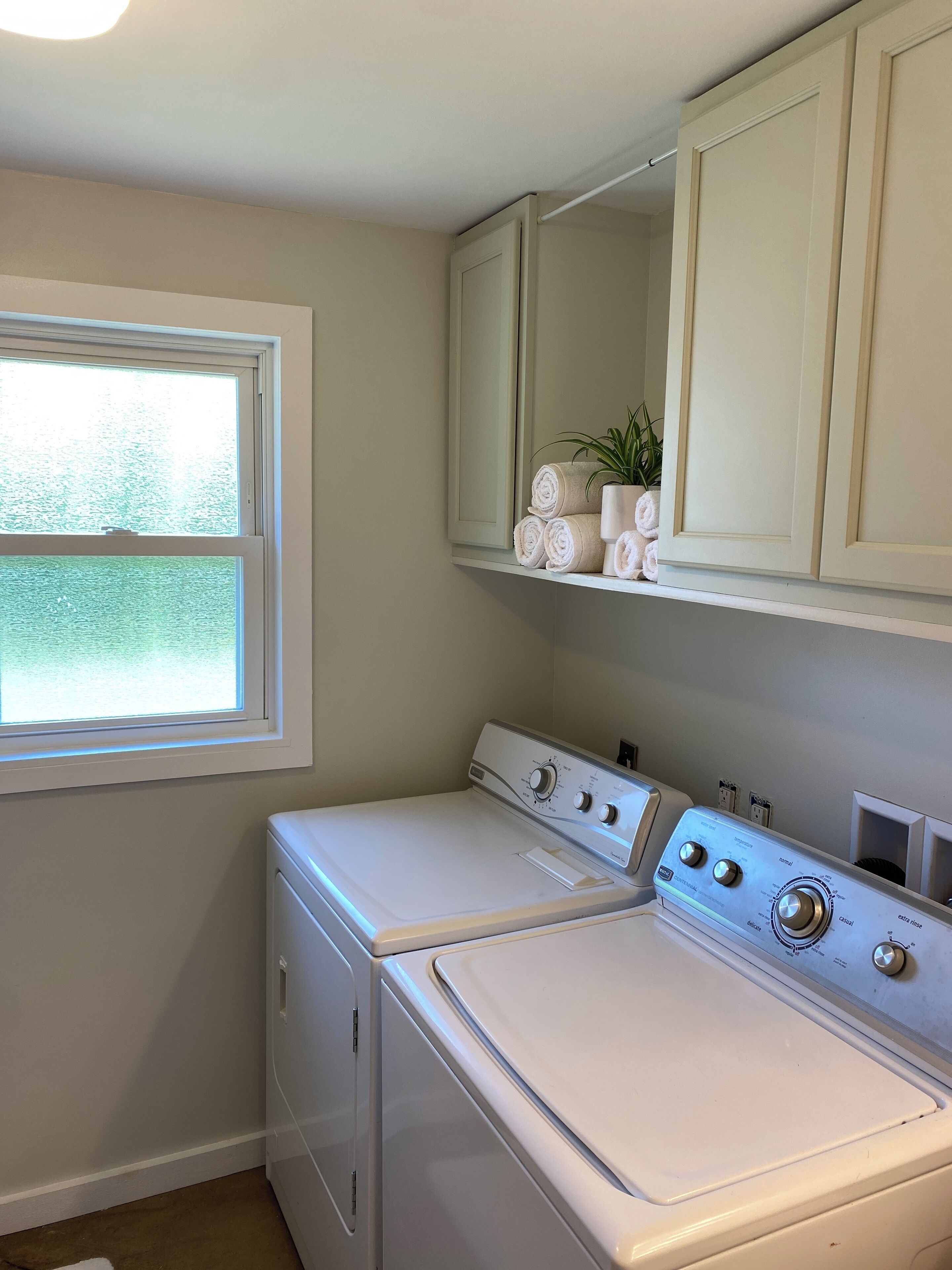 A laundry room with a front-loading washer and dryer, a window, and a shelf with towels and a plant.