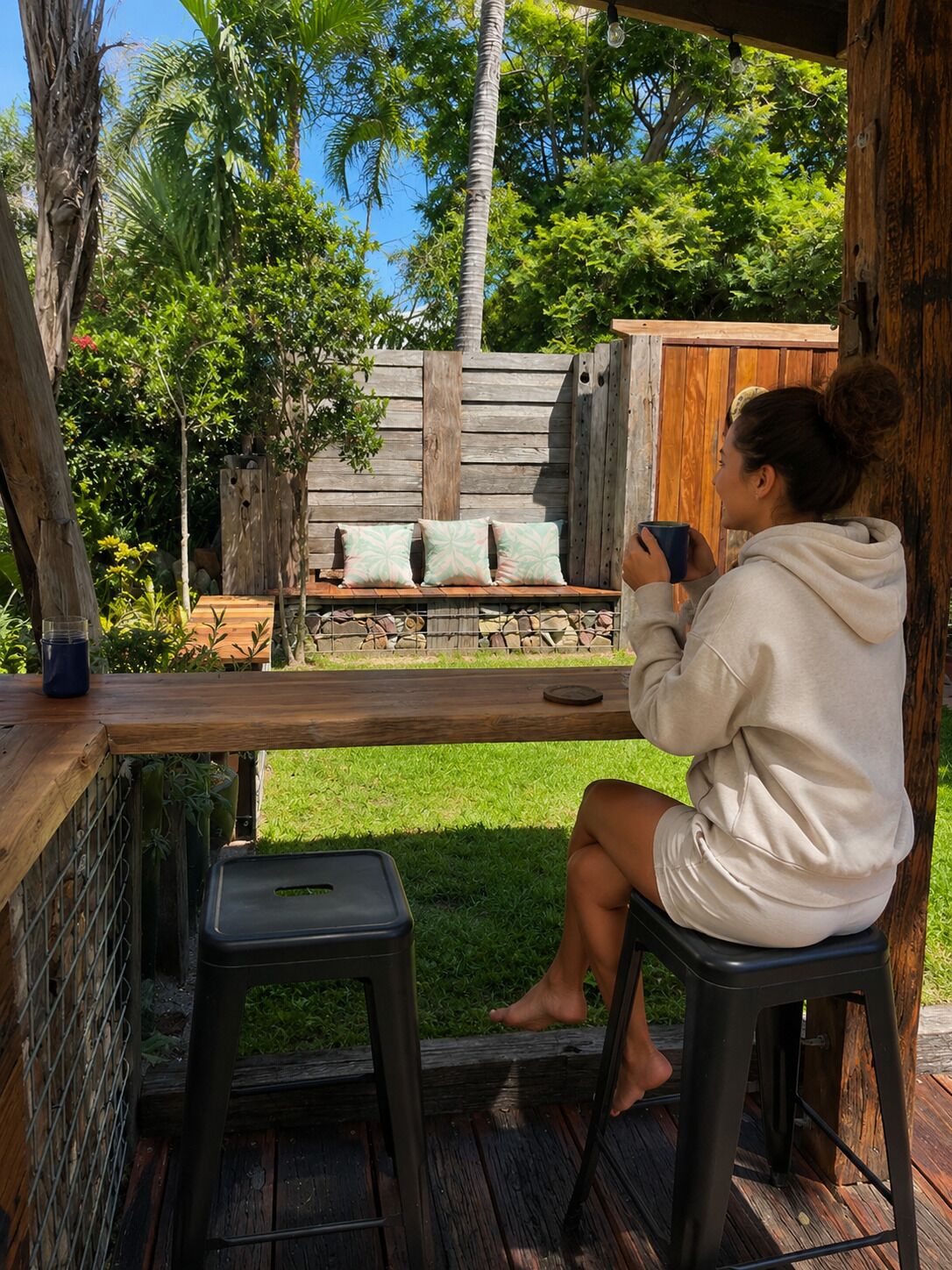 A person sitting on a stool at a wooden table, holding a mug, with a wooden bench and cushions in the background.
