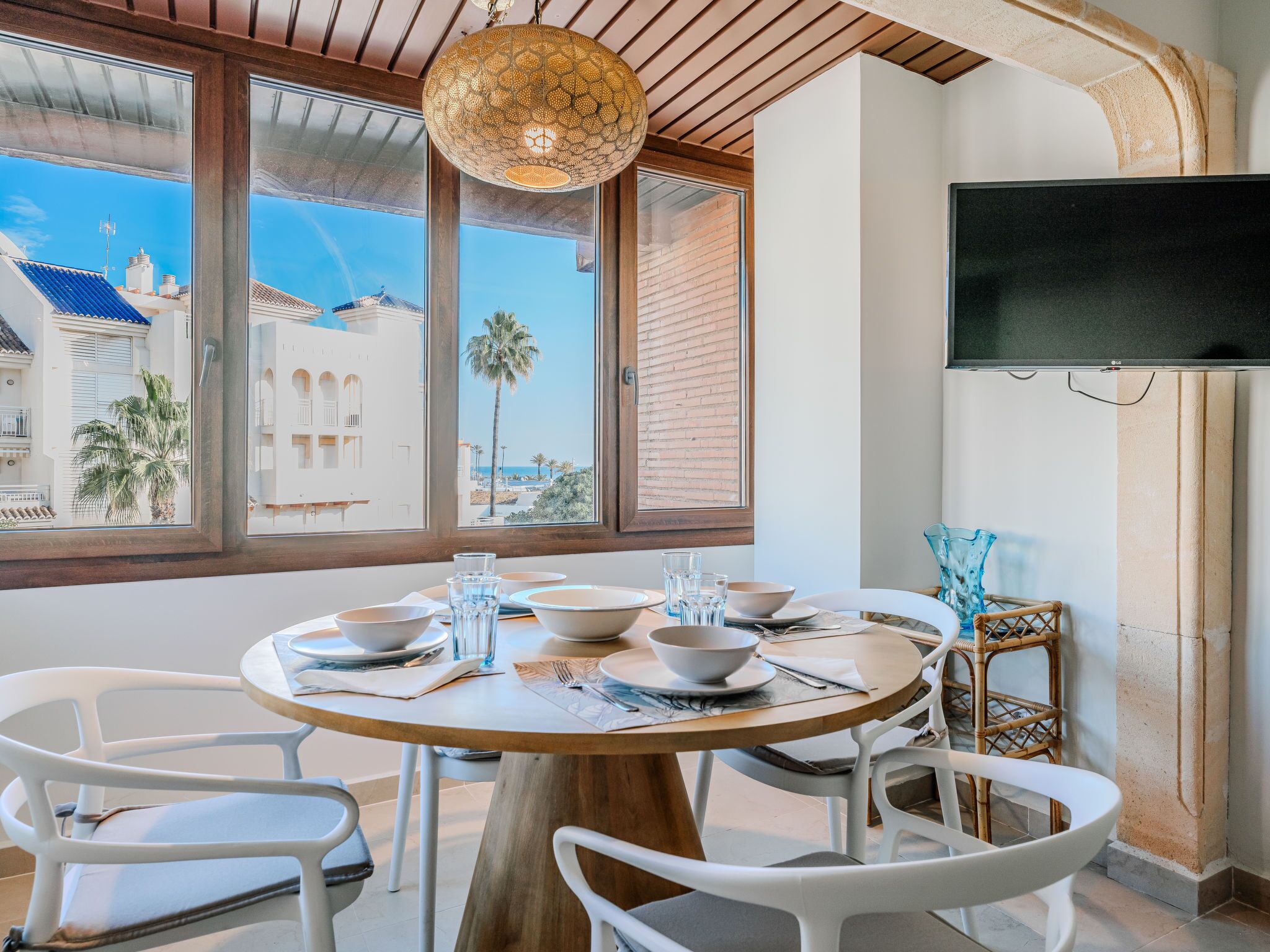 A dining area with a round wooden table, white chairs, and a large window with a view of buildings and palm trees.