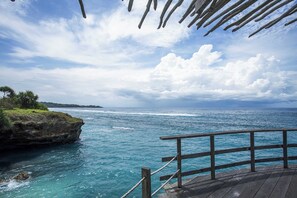 Un molo in legno con ringhiera, affacciato su un mare blu limpido e un litorale roccioso sotto un cielo parzialmente nuvoloso.