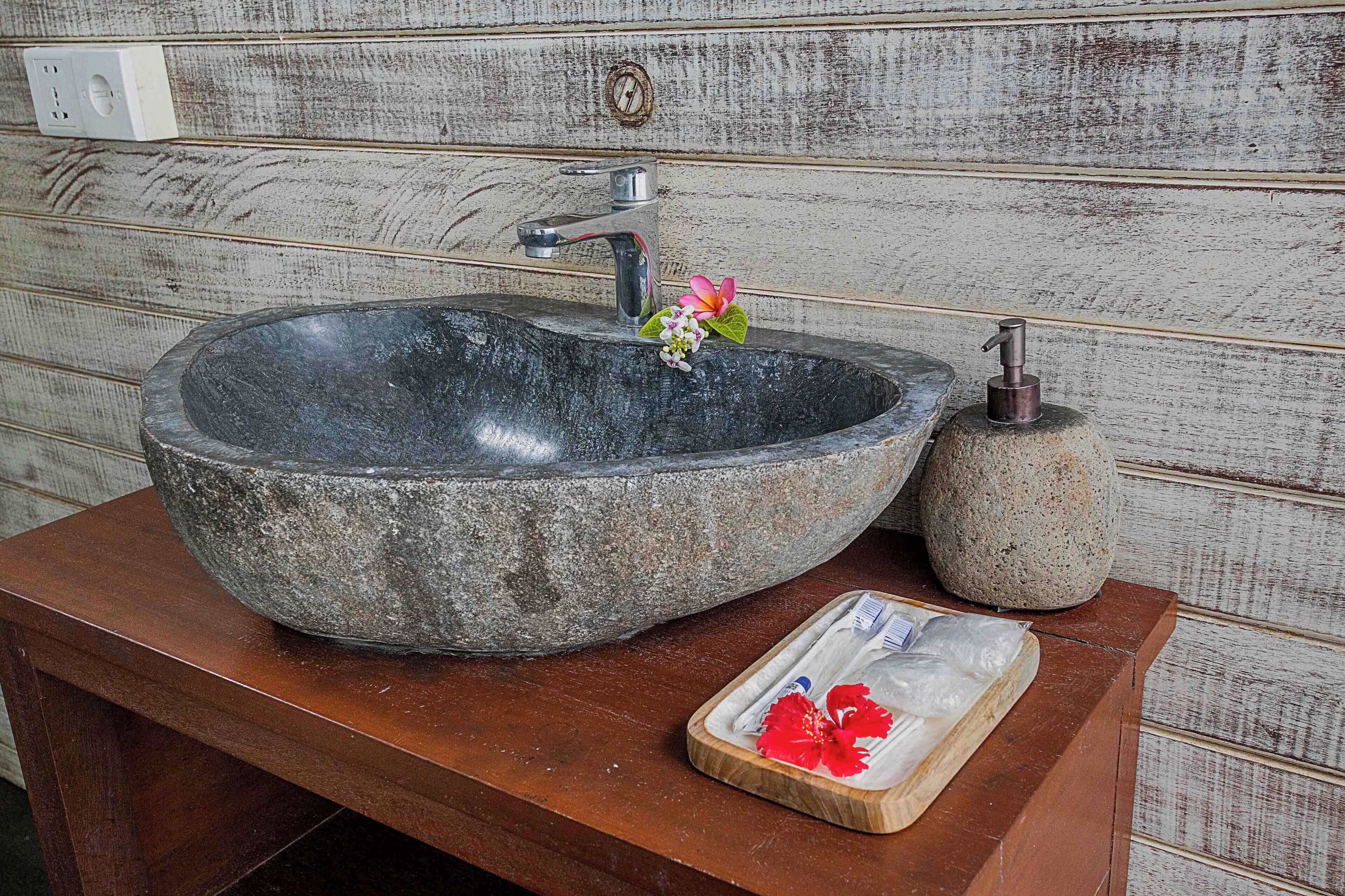 Een wastafel van natuursteen met een moderne kraan, een zeepdispenser en een houten dienblad met bloemen.