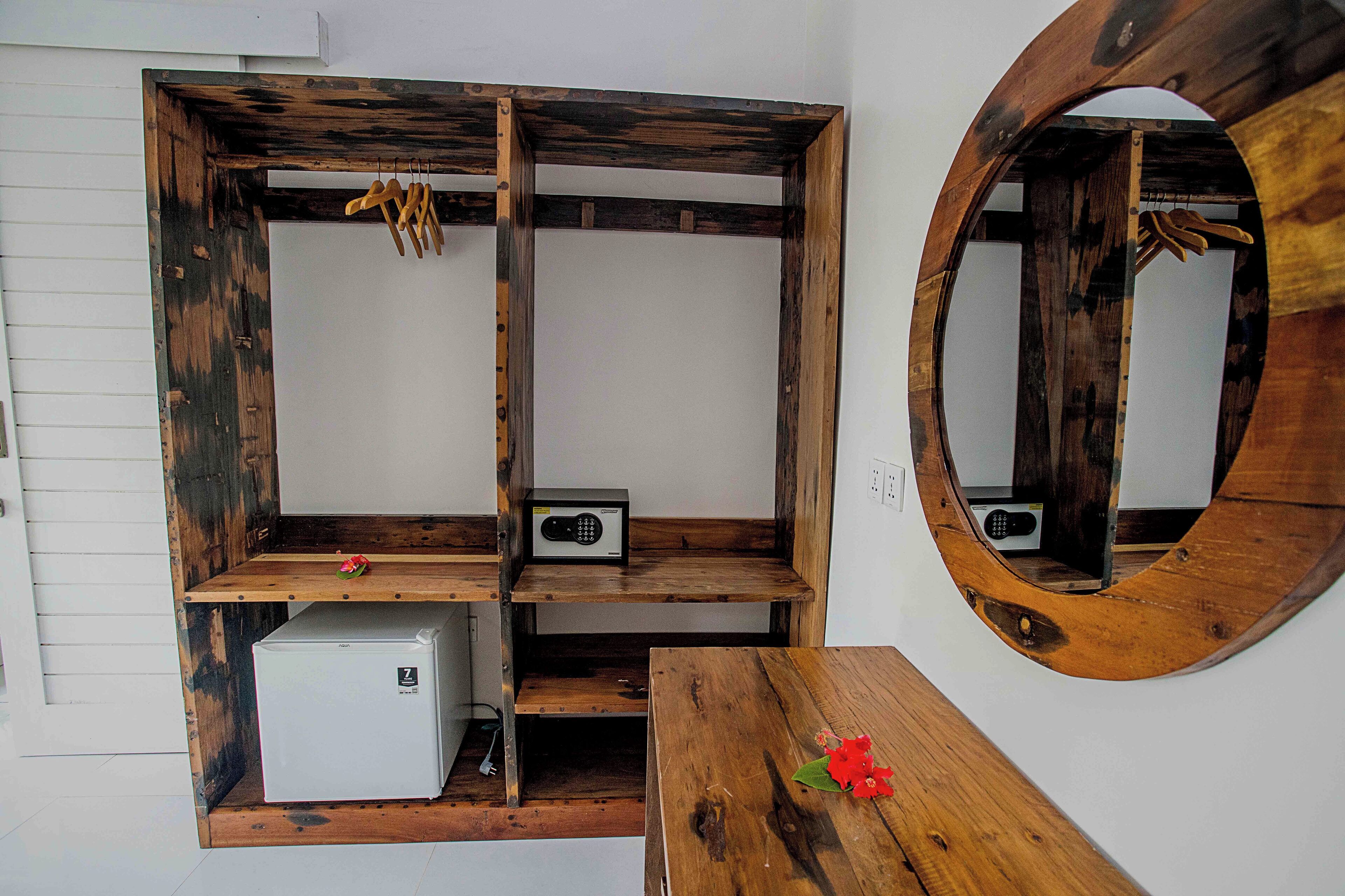 A room with a wooden wardrobe, a small white refrigerator, and a round mirror on the wall.